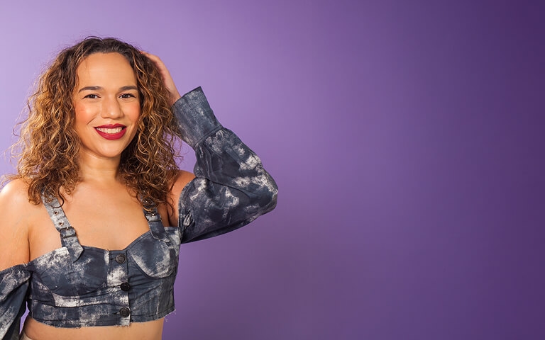 Confident person with curly hair smiling against a purple backdrop wearing a denim top