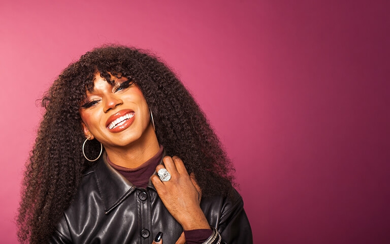 Person with curly hair and bold makeup smiling against a pink backdrop, wearing hoop earrings and a leather jacket