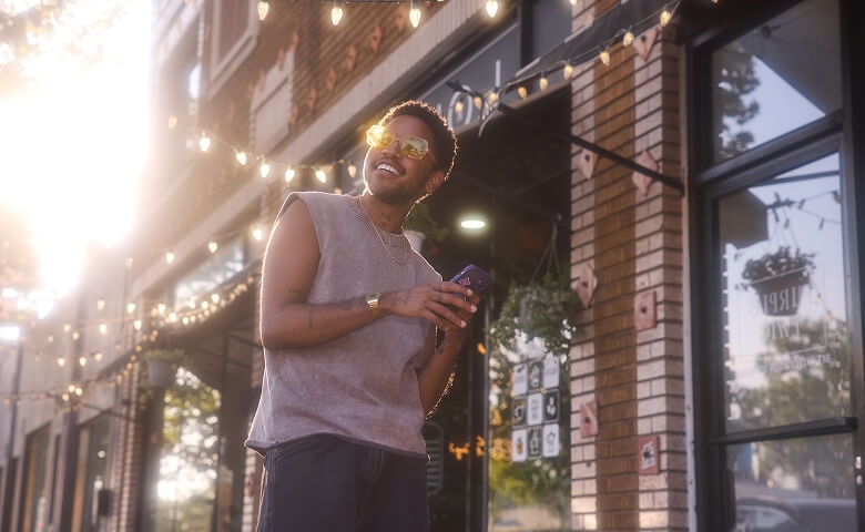 A stylish person wearing yellow-framed sunglasses, a sleeveless shirt, and a gold watch smiling while holding a smartphone outside a storefront during golden hour